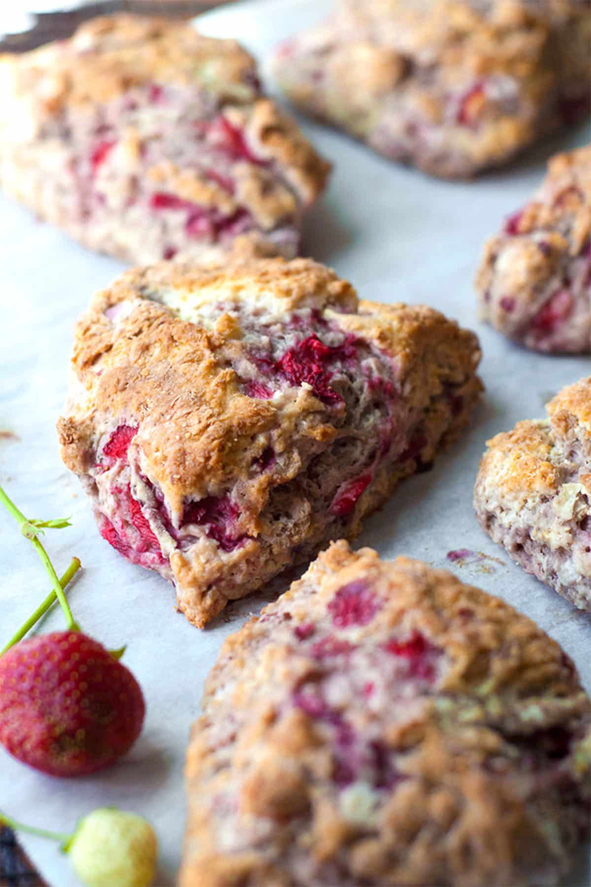 A close-up of fresh strawberry scones on parchment paper, with visible chunks of strawberries inside and a couple of whole strawberries nearby.