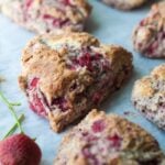 Fresh strawberry scones with visible pieces of strawberries are arranged on a sheet of parchment paper. Fresh strawberries are placed nearby for garnish.