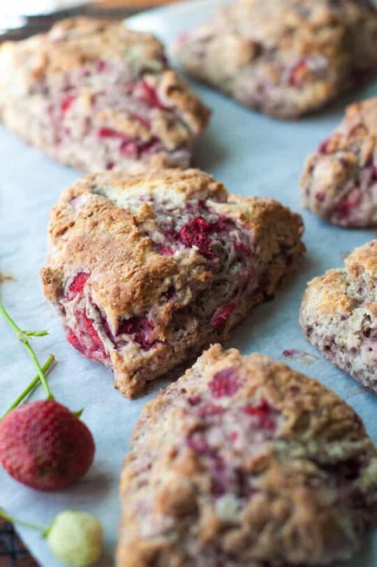 Fresh strawberry scones with visible pieces of strawberries are arranged on a sheet of parchment paper. Fresh strawberries are placed nearby for garnish.