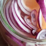 Close-up of pickled red onions submerged in a clear, slightly pink pickling liquid inside a glass jar. The onions appear fresh and vibrant, showcasing their crisp texture and tangy flavor.