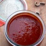 An open glass jar filled with thick, deep red easy enchilada sauce sits on a wooden surface. The jar lid and metal clasp are placed nearby.