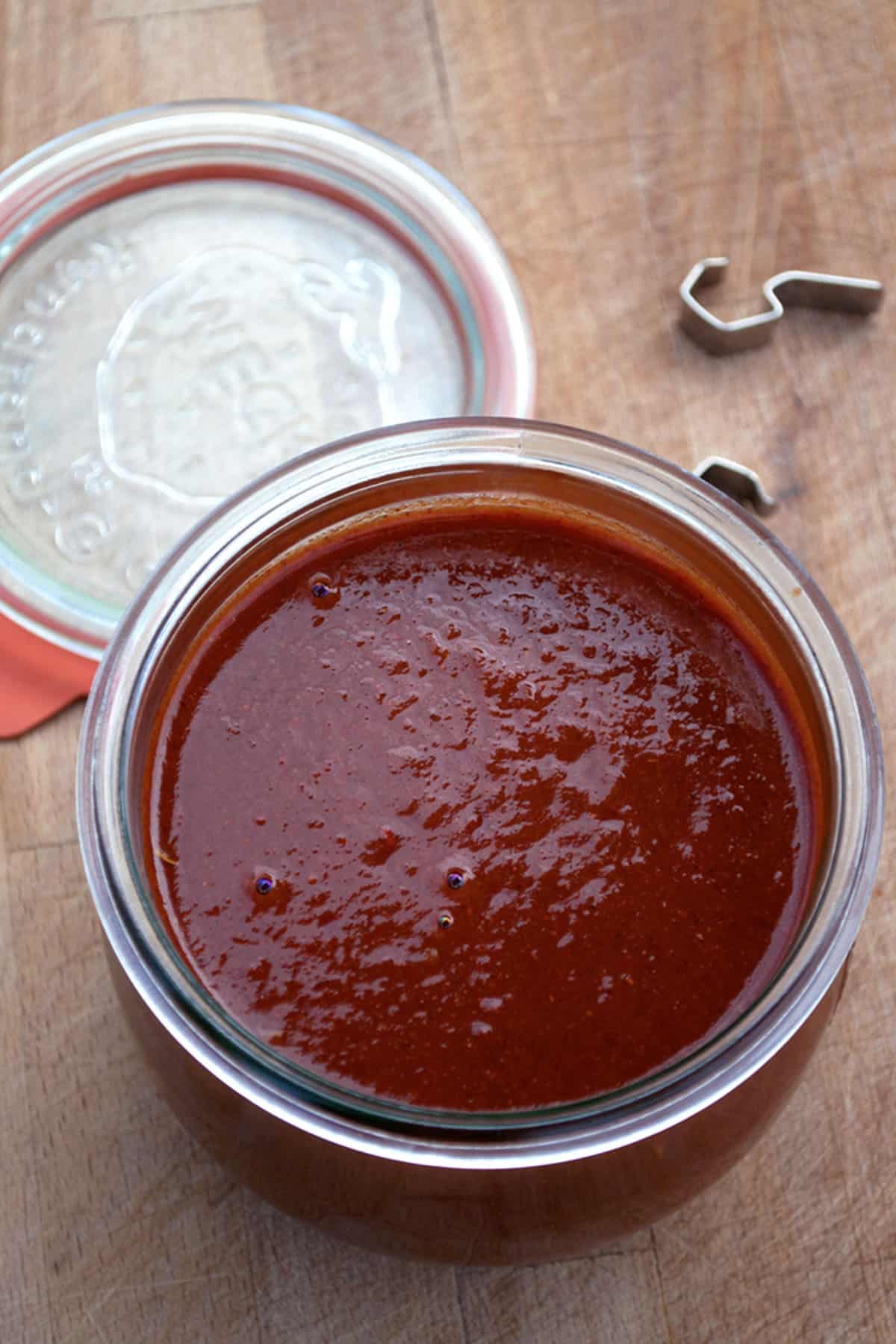 An open glass jar filled with thick, deep red easy enchilada sauce sits on a wooden surface. The jar lid and metal clasp are placed nearby.