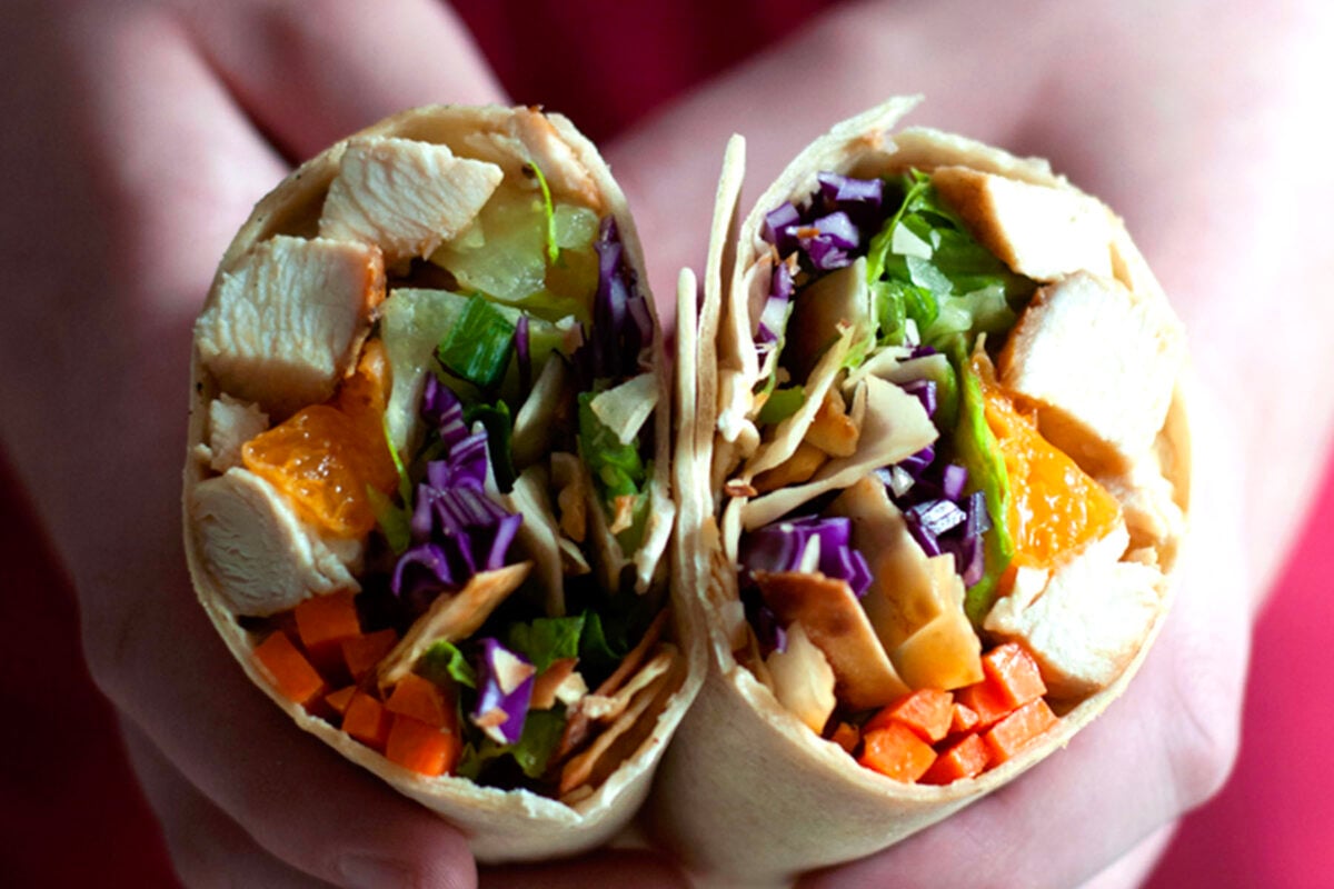 Two hands hold a wrap cut in half, revealing colorful fillings: diced chicken, lettuce, shredded purple cabbage, orange segments, and chopped carrots inside a tortilla—the perfect Asian chicken wraps for a fresh and flavorful meal.