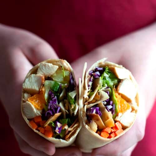 Two hands hold a sliced Chinese chicken wrap filled with tender chicken, lettuce, shredded purple cabbage, carrots, and other colorful vegetables. A red shirt is visible in the background.