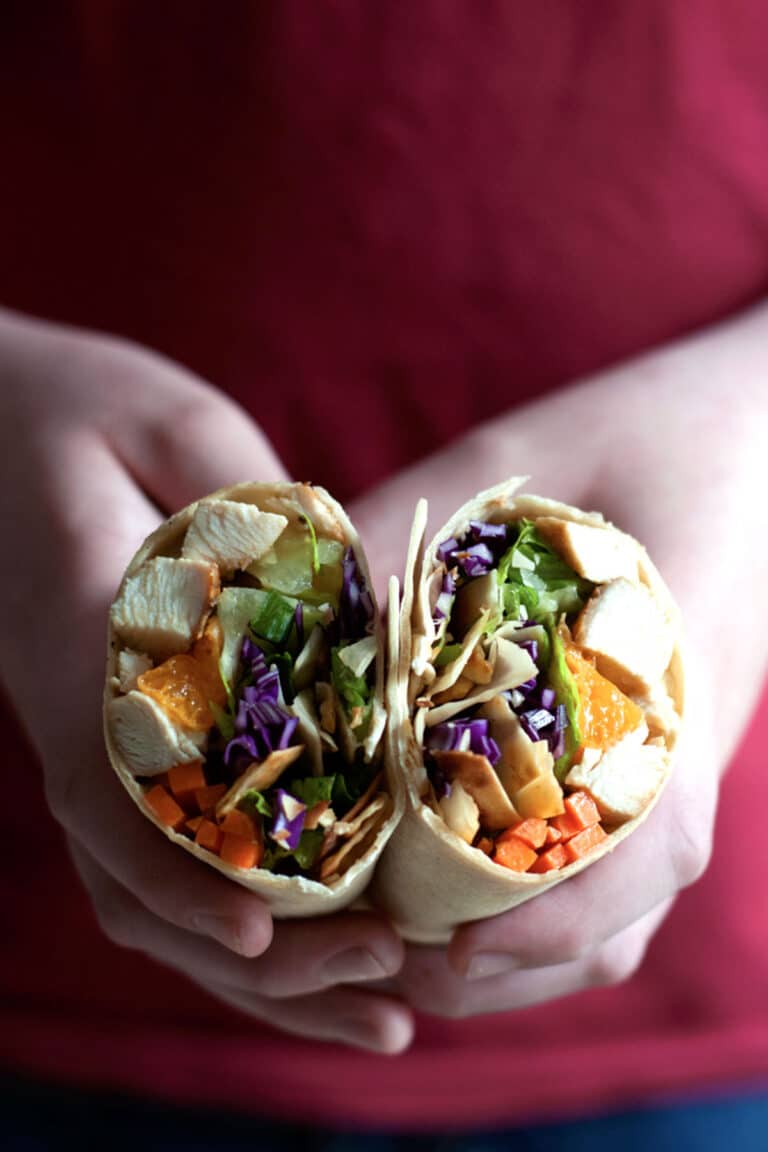 Two hands hold a sliced Chinese chicken wrap filled with tender chicken, lettuce, shredded purple cabbage, carrots, and other colorful vegetables. A red shirt is visible in the background.