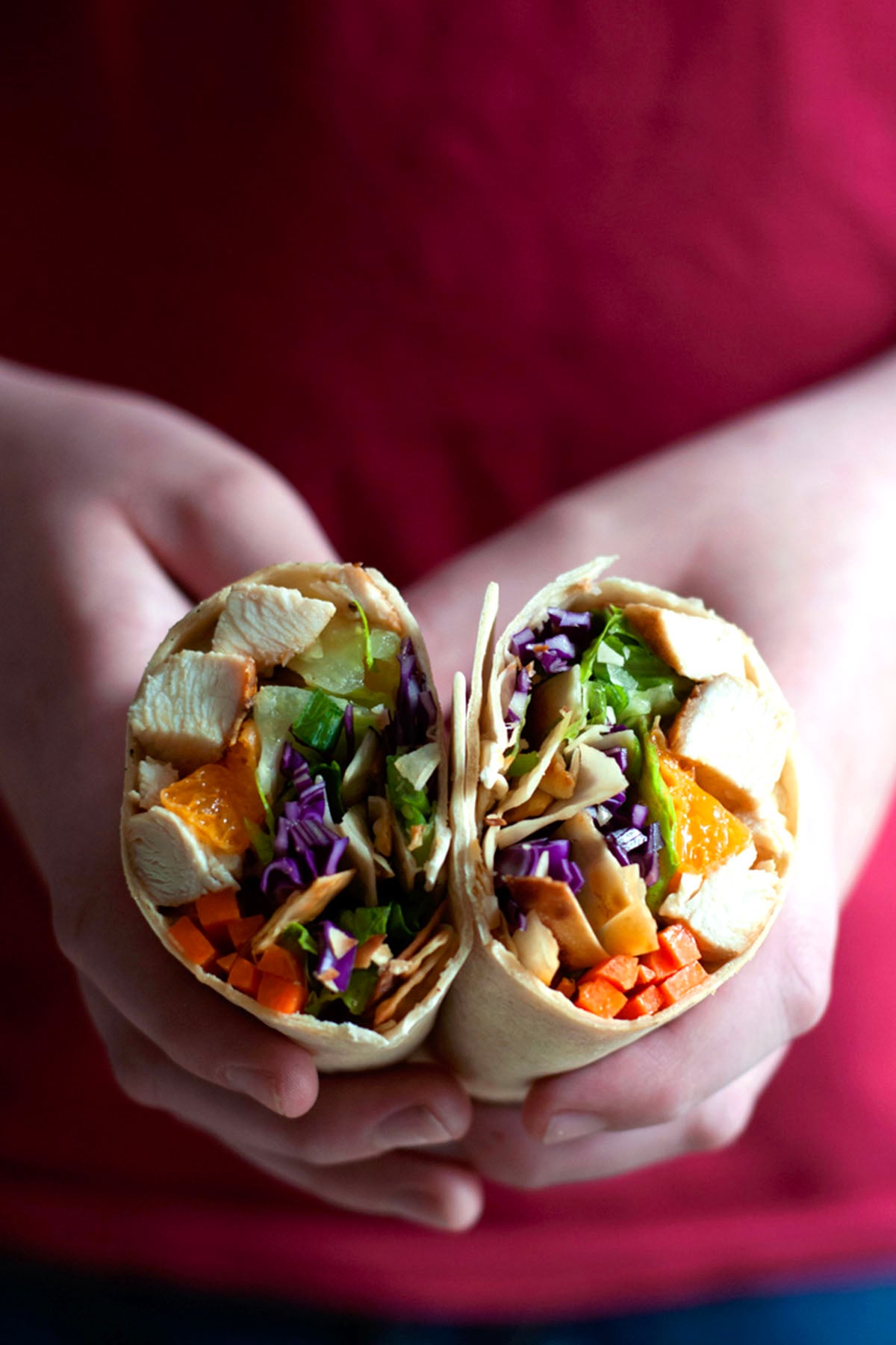 Two hands hold a sliced Chinese chicken wrap filled with tender chicken, lettuce, shredded purple cabbage, carrots, and other colorful vegetables. A red shirt is visible in the background.