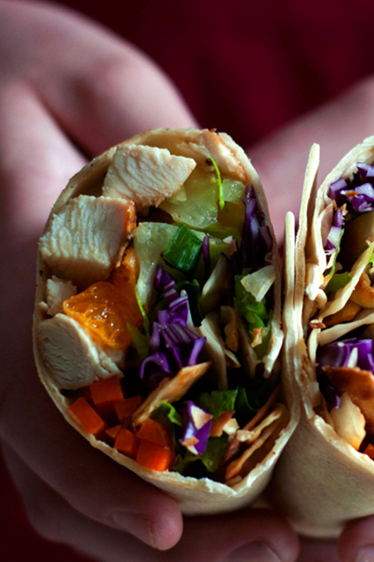 A close-up of a hand holding a wrap cut in half, showing colorful ingredients like chicken, purple cabbage, carrots, lettuce, and orange slices inside the tortilla—perfectly capturing the fresh flavors of Chinese chicken wraps.