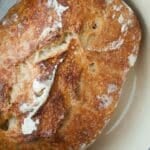 A round loaf of rustic, golden-brown no knead bread with a crisp crust, baked in a cream-colored Dutch oven.