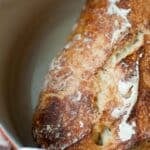 A close-up of a golden-brown, crusty loaf of no knead bread resting in a round, light-colored Dutch oven. Some patches of flour are visible on the surface of the artisanal bread.