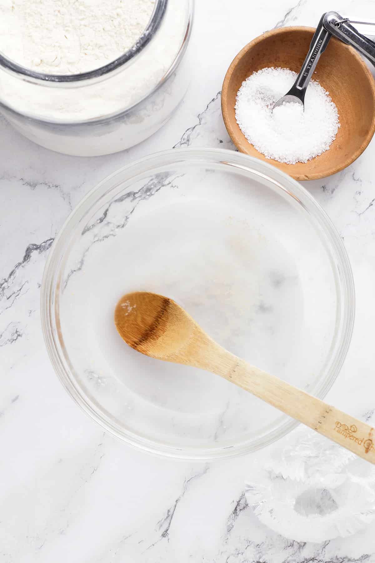 A glass bowl with water and a wooden spoon sits on a marble surface—essentials for no knead artisan bread. Nearby are a jar of flour and a small wooden bowl with salt and a measuring spoon, ready for your next overnight bread recipe.