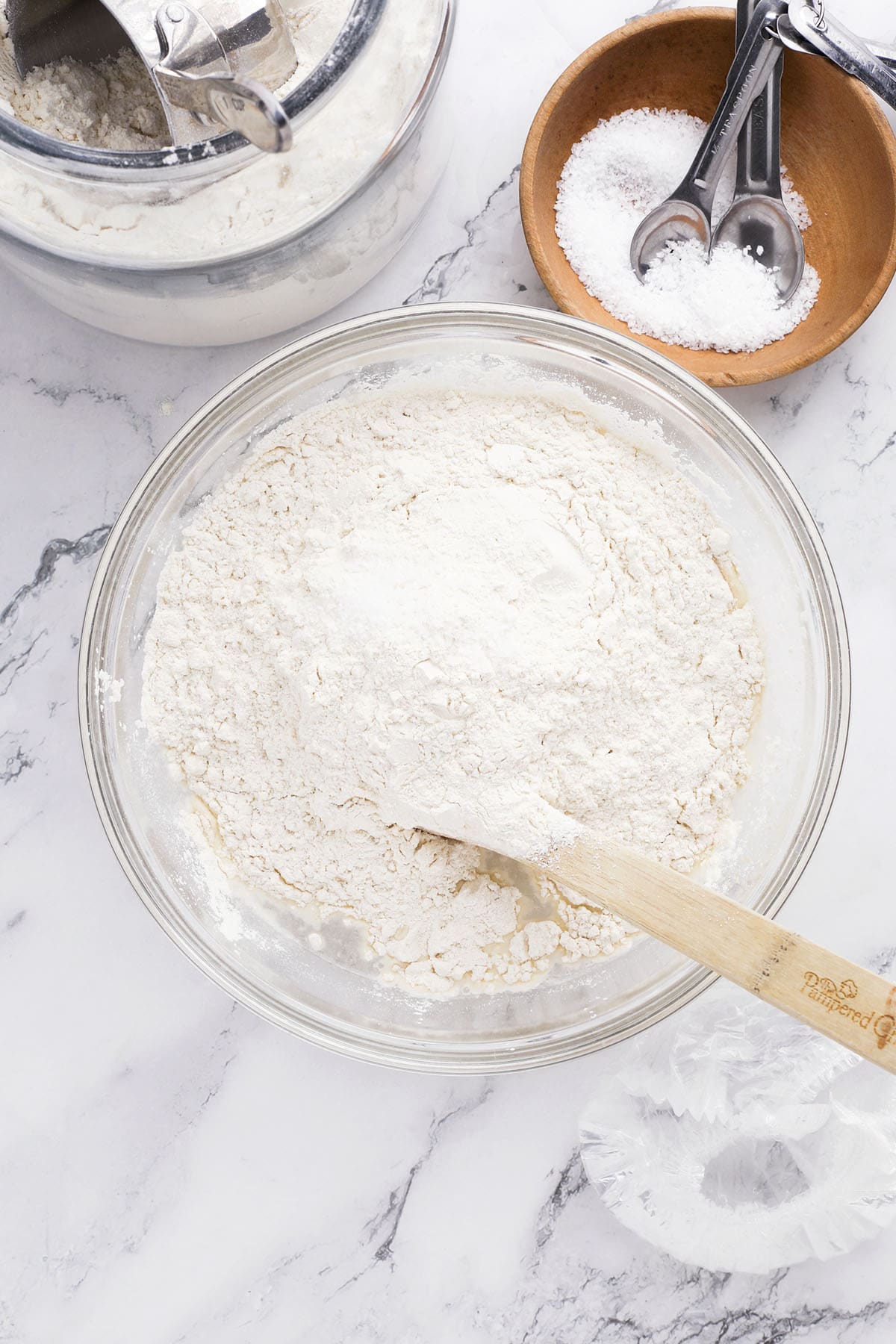 A glass bowl filled with flour and a wooden spoon rests on a marble countertop—perfect for preparing homemade artisan bread. Nearby are a container of flour with a scoop and a small wooden bowl holding salt and measuring spoons.