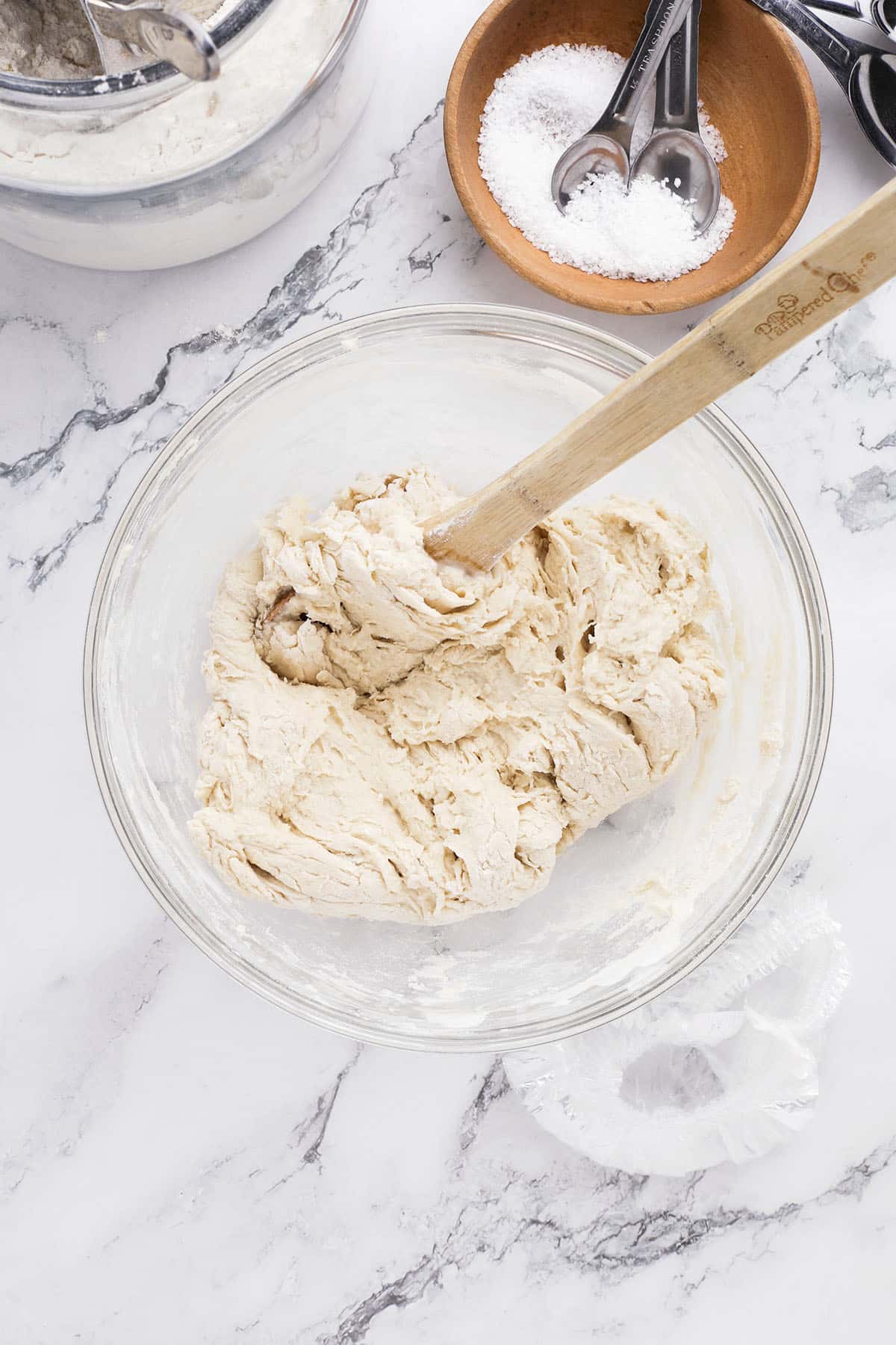 A glass bowl with no knead bread dough being mixed by a wooden spatula sits on a marble countertop. Nearby are a bowl of salt with measuring spoons and a metal bowl filled with flour, ready for your next dutch oven bread creation.