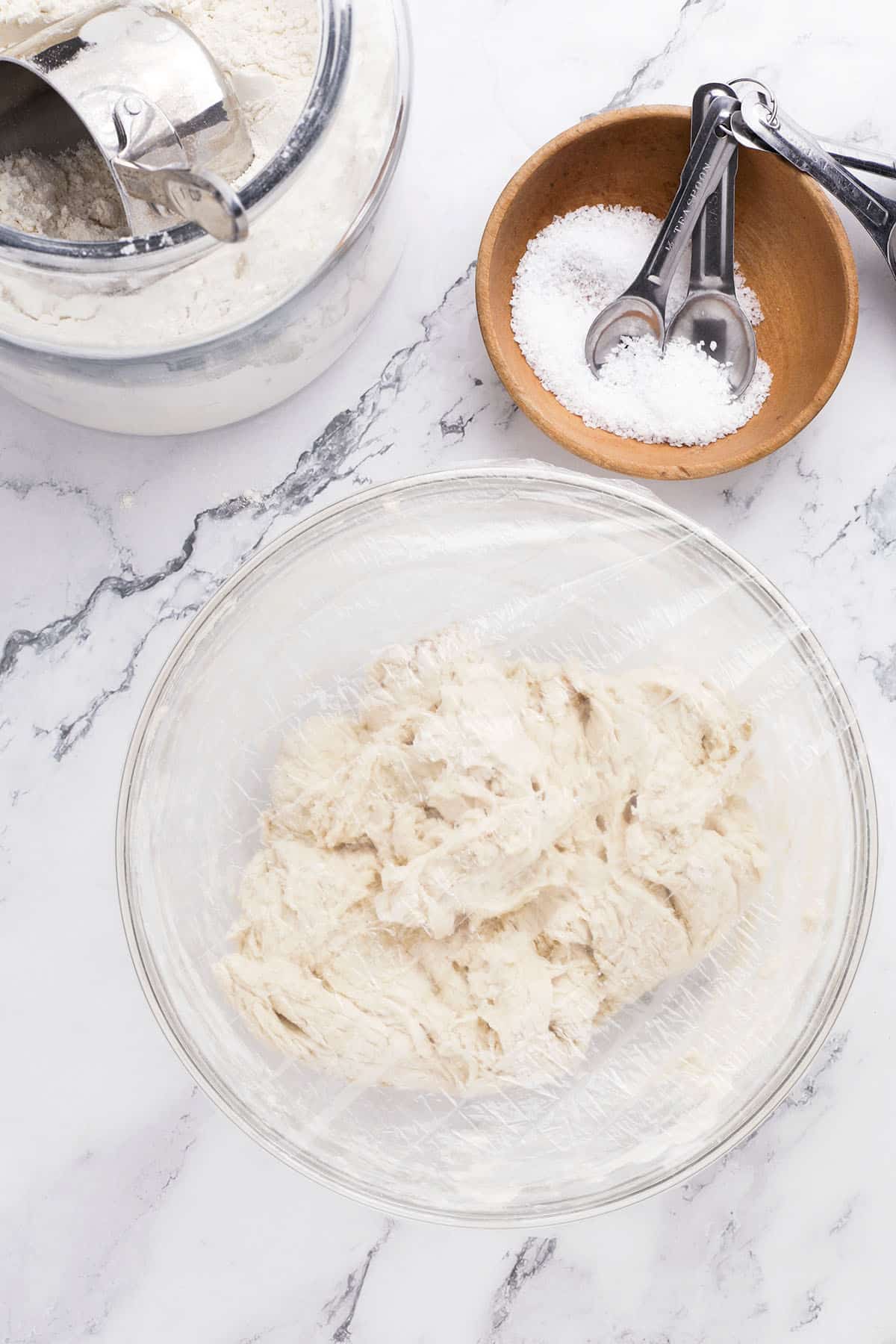 A glass bowl with no knead bread dough covered in plastic wrap sits on a marble counter. Nearby are a wooden bowl with salt and measuring spoons, and a jar of flour with a metal scoop—perfect for homemade artisan bread.