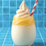A glass of creamy, swirled Dole Whip pineapple soft serve topped with a red and white striped straw, set against a bright blue tiled background.