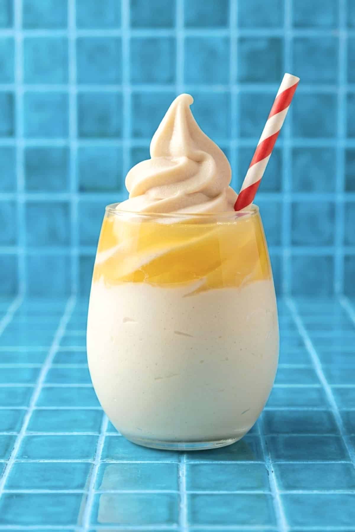 A glass of creamy, swirled Dole Whip pineapple soft serve topped with a red and white striped straw, set against a bright blue tiled background.