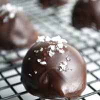 A close-up of a round dark chocolate chile truffle topped with flaky sea salt, resting on a black wire cooling rack with other truffles blurred in the background.