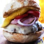 A close-up of a Cuban burger on a rustic bun with melted cheese, red onion slices, tomato, and a pickle spear, sitting on a wooden surface.