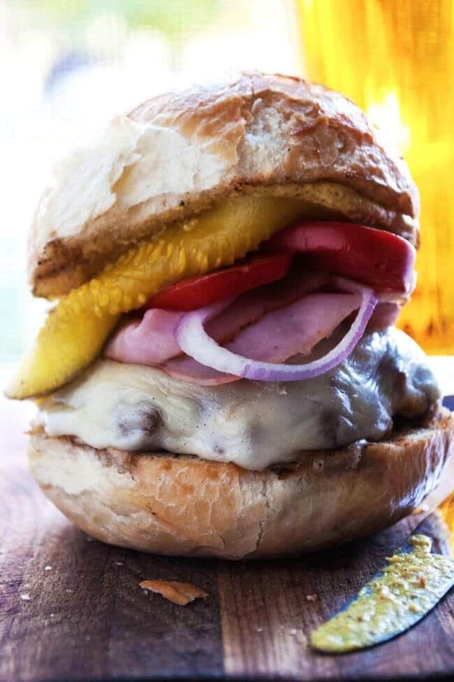 A close-up of a Cuban burger on a rustic bun with melted cheese, red onion slices, tomato, and a pickle spear, sitting on a wooden surface.
