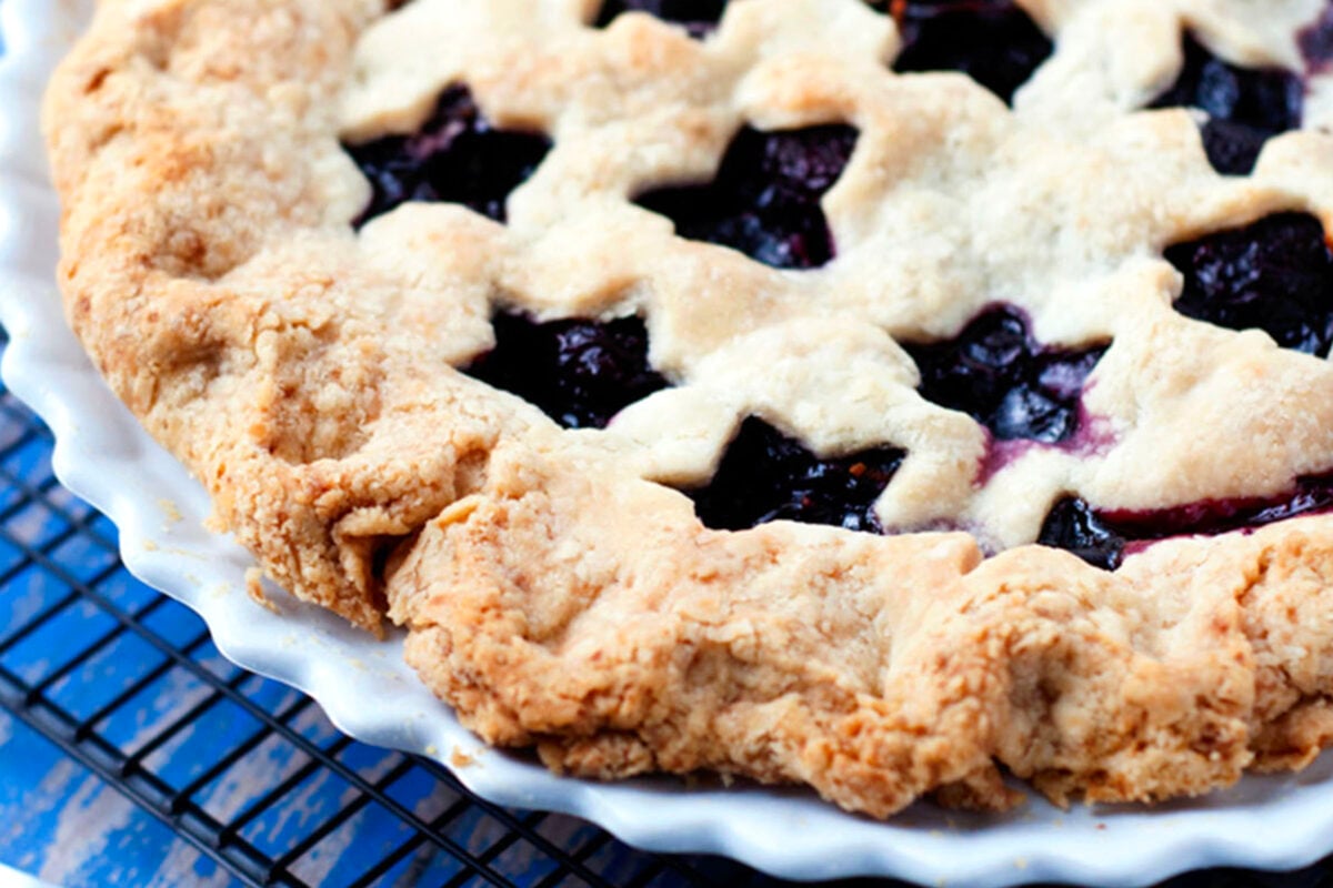 A close-up of a freshly baked cherry pie with a golden, flaky lattice crust, filled with dark berry and cherry filling, in a white pie dish on a cooling rack.