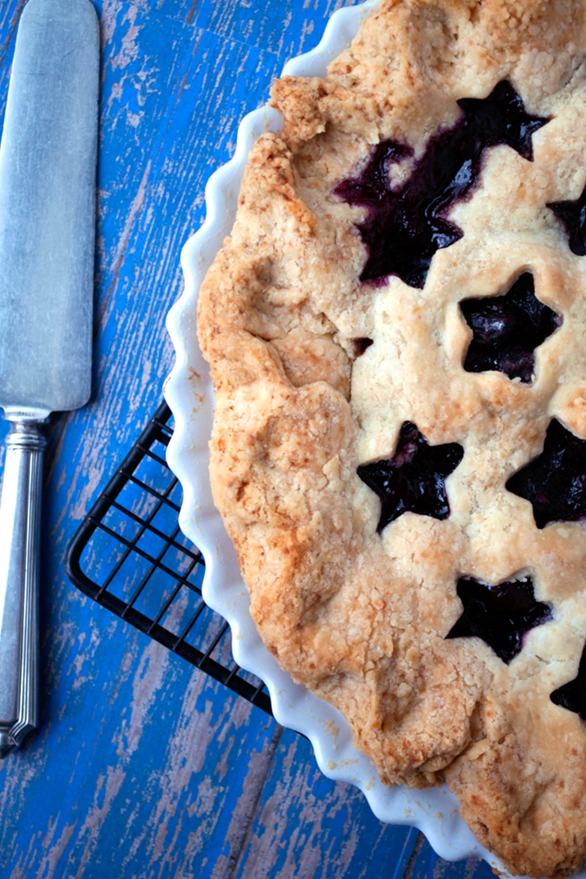 A close-up of a cherry pie with a golden, flaky crust and star-shaped cutouts revealing a dark berry filling, sitting in a white pie dish on a cooling rack, next to a pie server on a blue wooden surface.