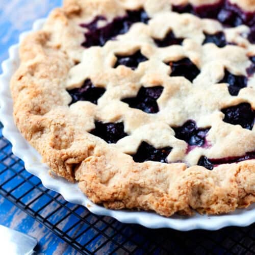 A homemade cherry pie with a star-shaped lattice crust sits in a white pie dish on a cooling rack, next to a pie server on a blue wooden surface.