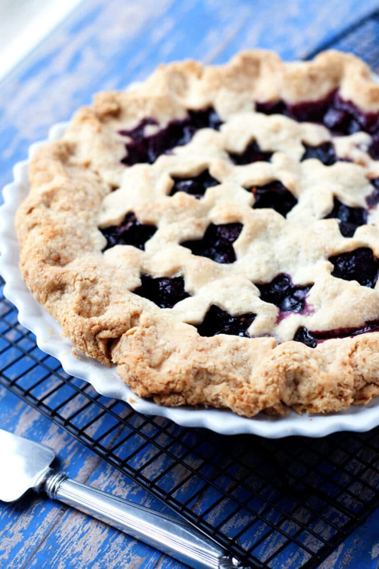 A homemade cherry pie with a star-shaped lattice crust sits in a white pie dish on a cooling rack, next to a pie server on a blue wooden surface.
