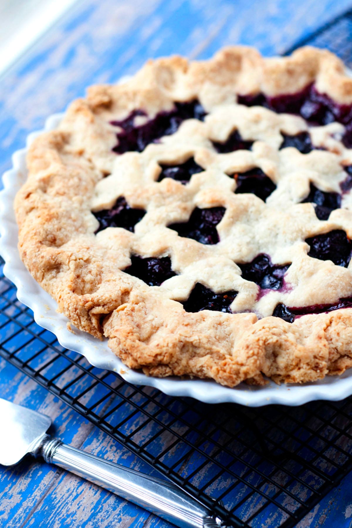 A homemade cherry pie with a star-shaped lattice crust sits in a white pie dish on a cooling rack, next to a pie server on a blue wooden surface.