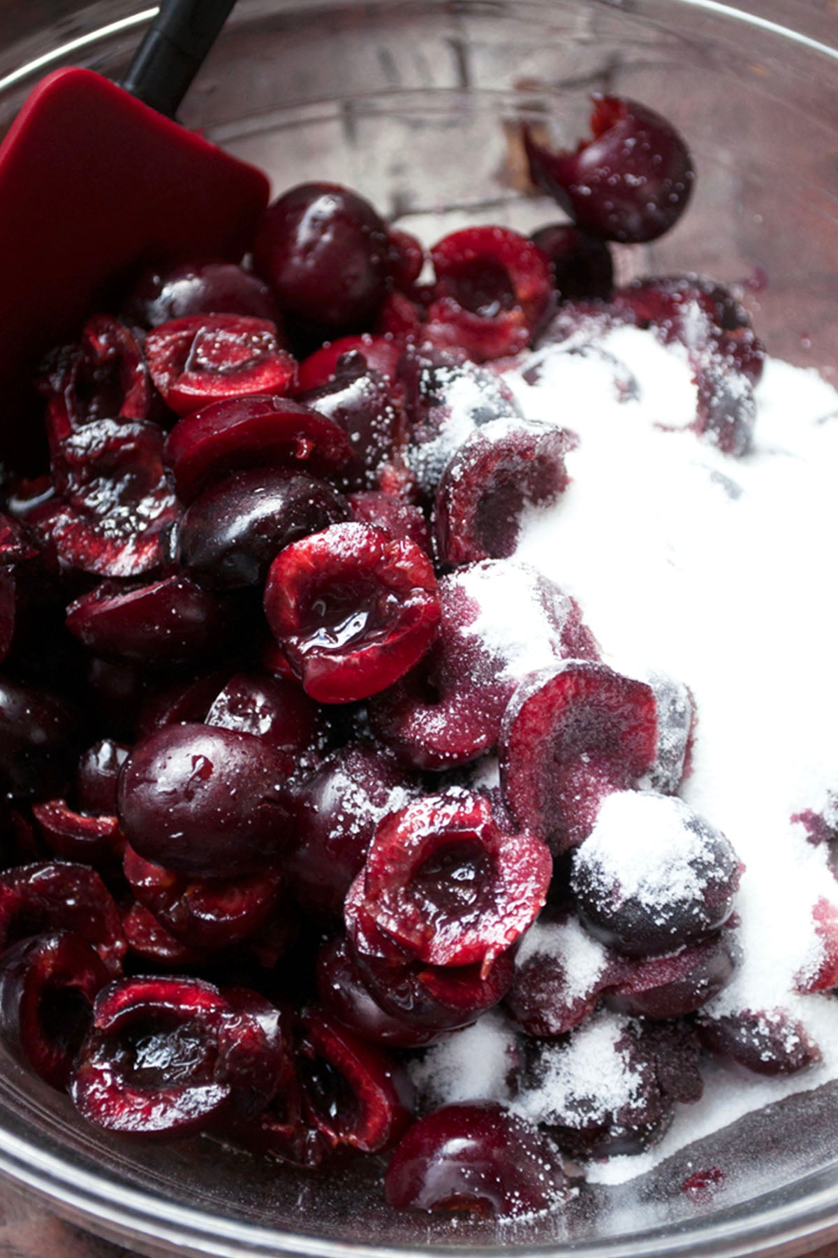 A close-up of a glass bowl filled with halved dark cherries and granulated sugar, perfect for making cherry pie, with a red spatula partially visible in the top left corner.