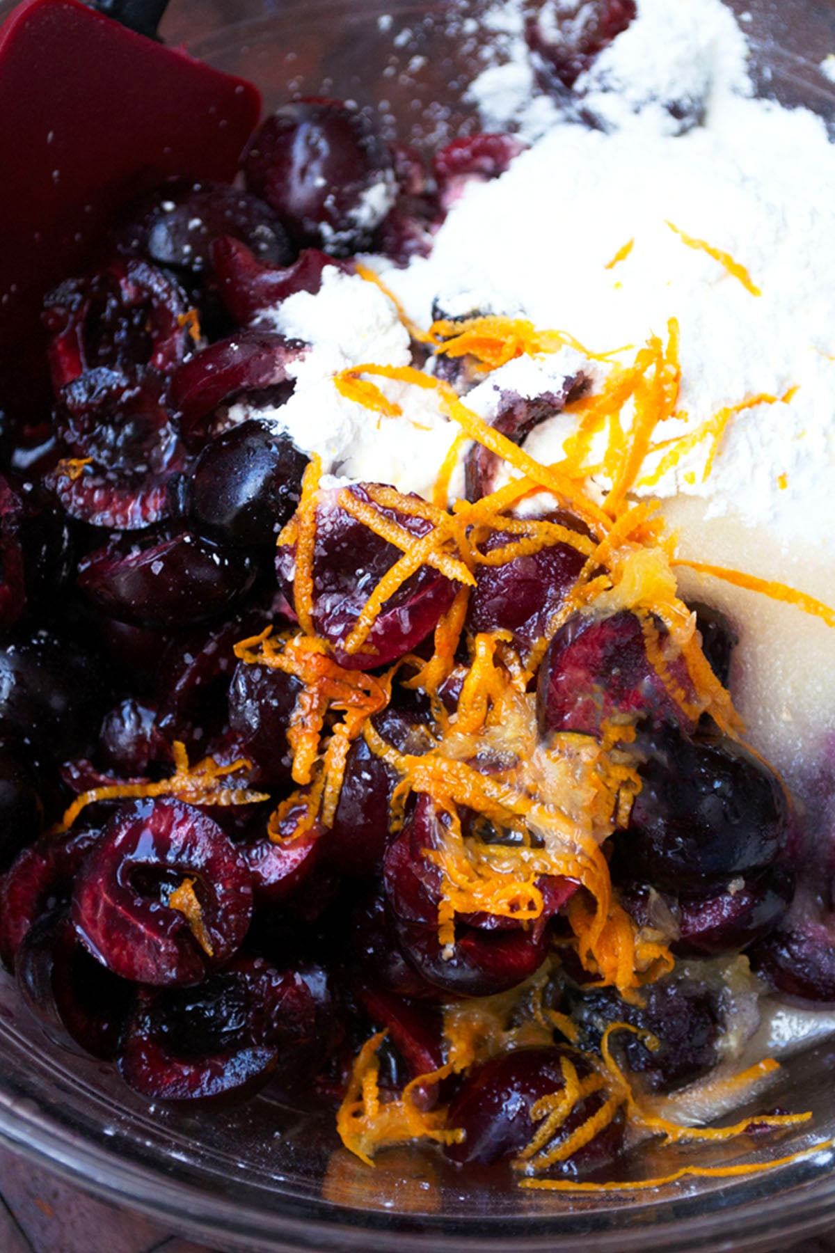 A close-up of a bowl containing halved dark cherries, grated orange zest, white sugar, and flour—classic ingredients for a cherry pie—with a red spatula partially visible on the side.