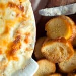 A baking dish filled with cheesy, browned artichoke dip sits next to a wooden board topped with toasted baguette slices and a knife with some spread on it.
