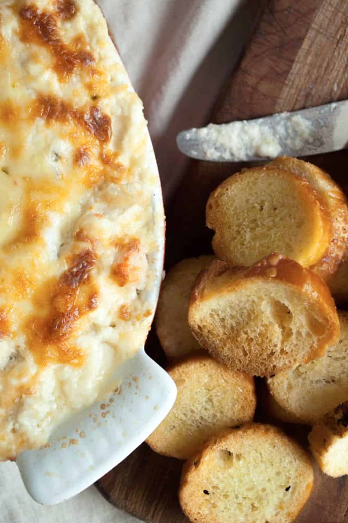 A baking dish filled with cheesy, browned artichoke dip sits next to a wooden board topped with toasted baguette slices and a knife with some spread on it.