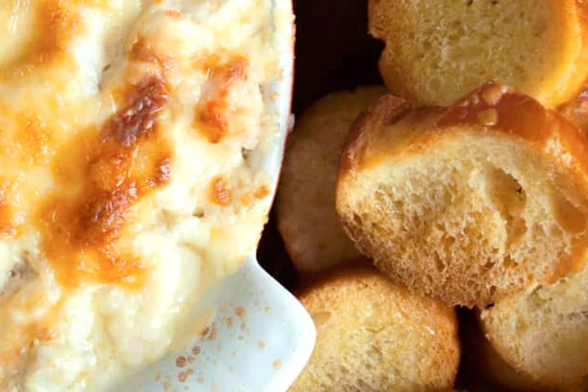 A close-up of cheesy baked artichoke dip with cream cheese in a white dish next to several slices of toasted baguette—a warm artichoke dip appetizer that's perfect as an easy party dip recipe.
