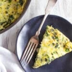 A slice of spinach artichoke frittata with cheese sits on a dark plate with a fork, beside a pan holding the rest of the frittata, all arranged on a light wooden surface.