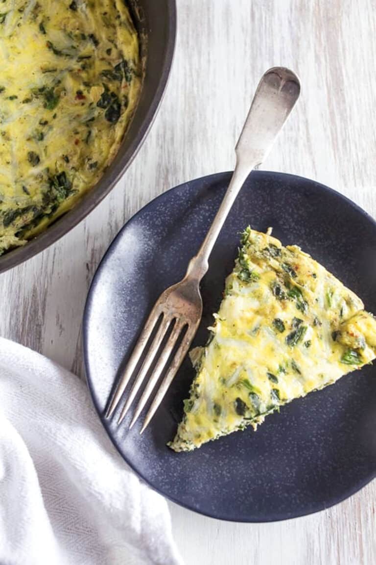 A slice of spinach artichoke frittata with cheese sits on a dark plate with a fork, beside a pan holding the rest of the frittata, all arranged on a light wooden surface.
