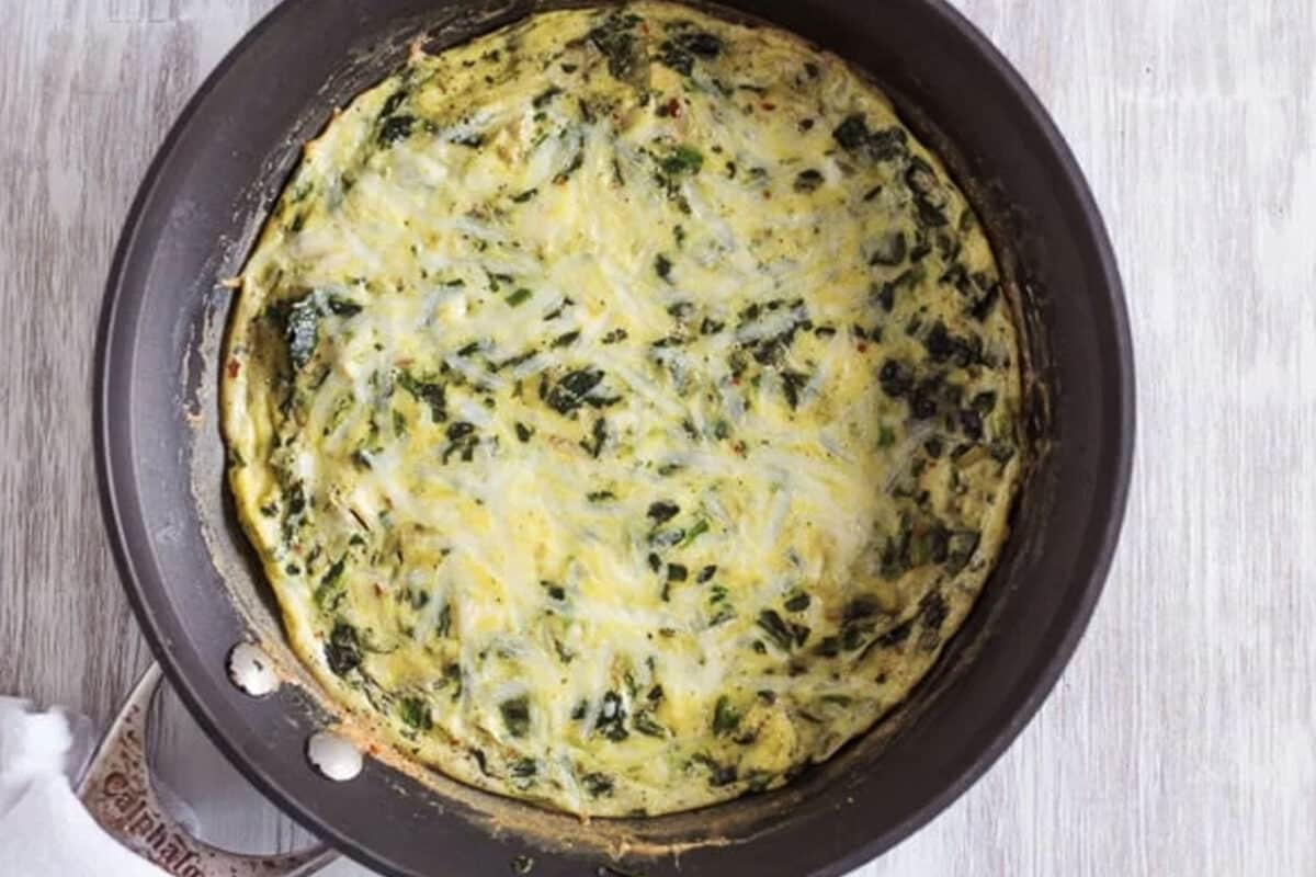 A cooked spinach artichoke frittata sits in a nonstick skillet on a light wooden surface, showcasing a golden, slightly browned edge and a mixture of green spinach, tender artichokes, and melted cheese.