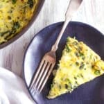 A slice of baked spinach artichoke frittata rests on a dark plate with a fork, next to the skillet frittata containing the rest of this delicious dish, all arranged on a light wooden surface—perfect for a hearty frittata for brunch.