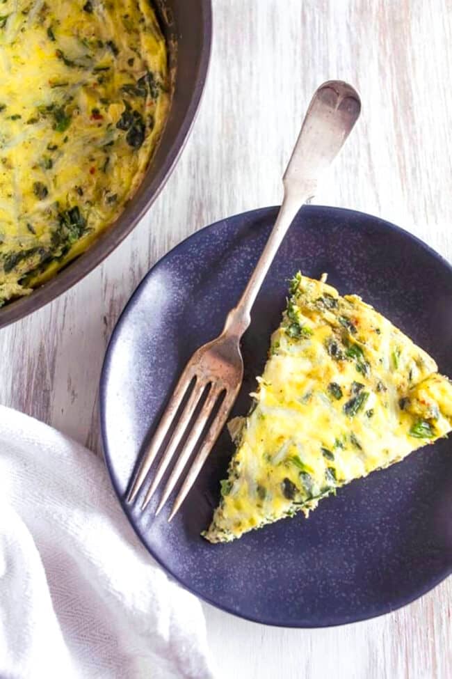 A slice of baked spinach artichoke frittata rests on a dark plate with a fork, next to the skillet frittata containing the rest of this delicious dish, all arranged on a light wooden surface—perfect for a hearty frittata for brunch.