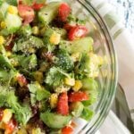 A glass bowl filled with a colorful corn avocado cucumber salad, featuring cherry tomatoes, cilantro, and black pepper, placed on a white and green striped cloth.