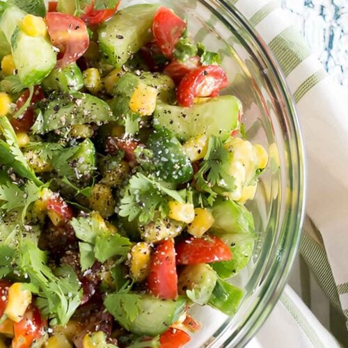 A glass bowl filled with a colorful corn avocado cucumber salad, featuring cherry tomatoes, cilantro, and black pepper, placed on a white and green striped cloth.