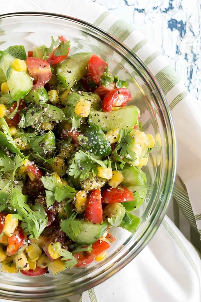 A glass bowl filled with a colorful corn avocado cucumber salad, featuring cherry tomatoes, cilantro, and black pepper, placed on a white and green striped cloth.