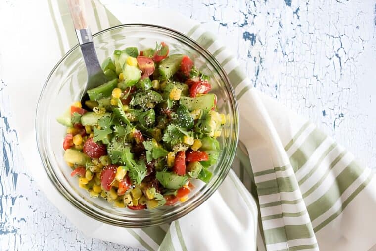 A glass bowl filled with a colorful corn avocado cucumber salad, featuring tomatoes and cilantro, sits on a white and green striped cloth on a textured white surface. A serving spoon rests in the bowl.