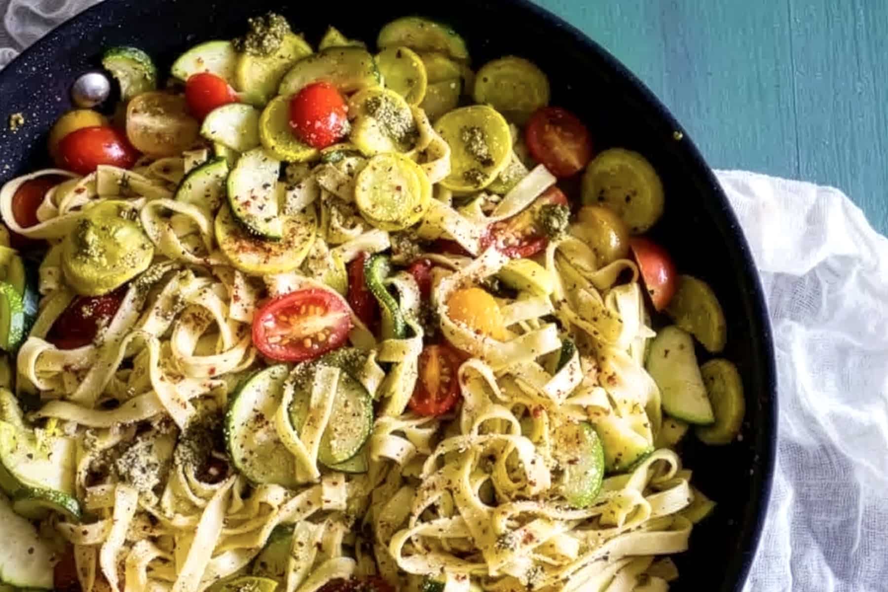 Tagliatelle pasta in a black pan with sliced zucchini, yellow squash, and halved cherry tomatoes on a teal surface. This colorful summer vegetable pesto pasta is sprinkled with herbs and seasonings. A white cloth is partially visible beside the pan.