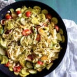 A bowl of summer vegetable pesto pasta, featuring fettuccine mixed with yellow squash, zucchini, and cherry tomatoes, topped with herbs, sits on a green table with a white cloth underneath.
