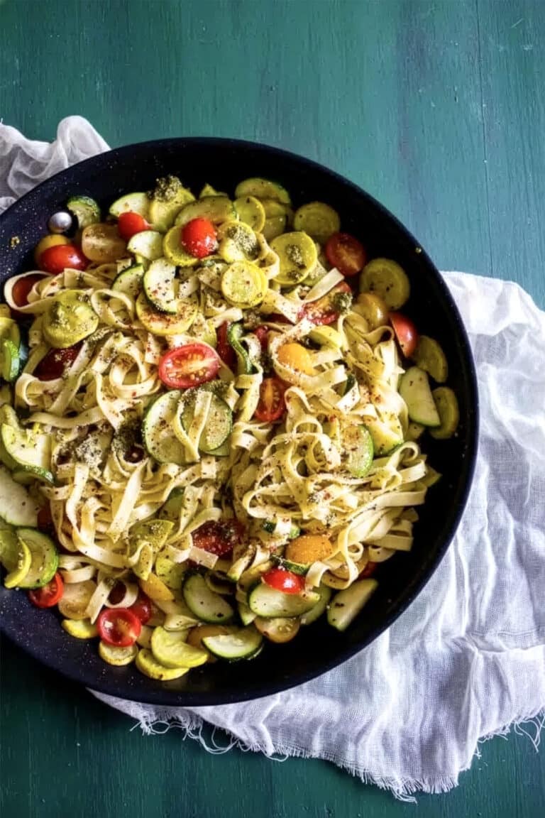 A bowl of summer vegetable pesto pasta, featuring fettuccine mixed with yellow squash, zucchini, and cherry tomatoes, topped with herbs, sits on a green table with a white cloth underneath.