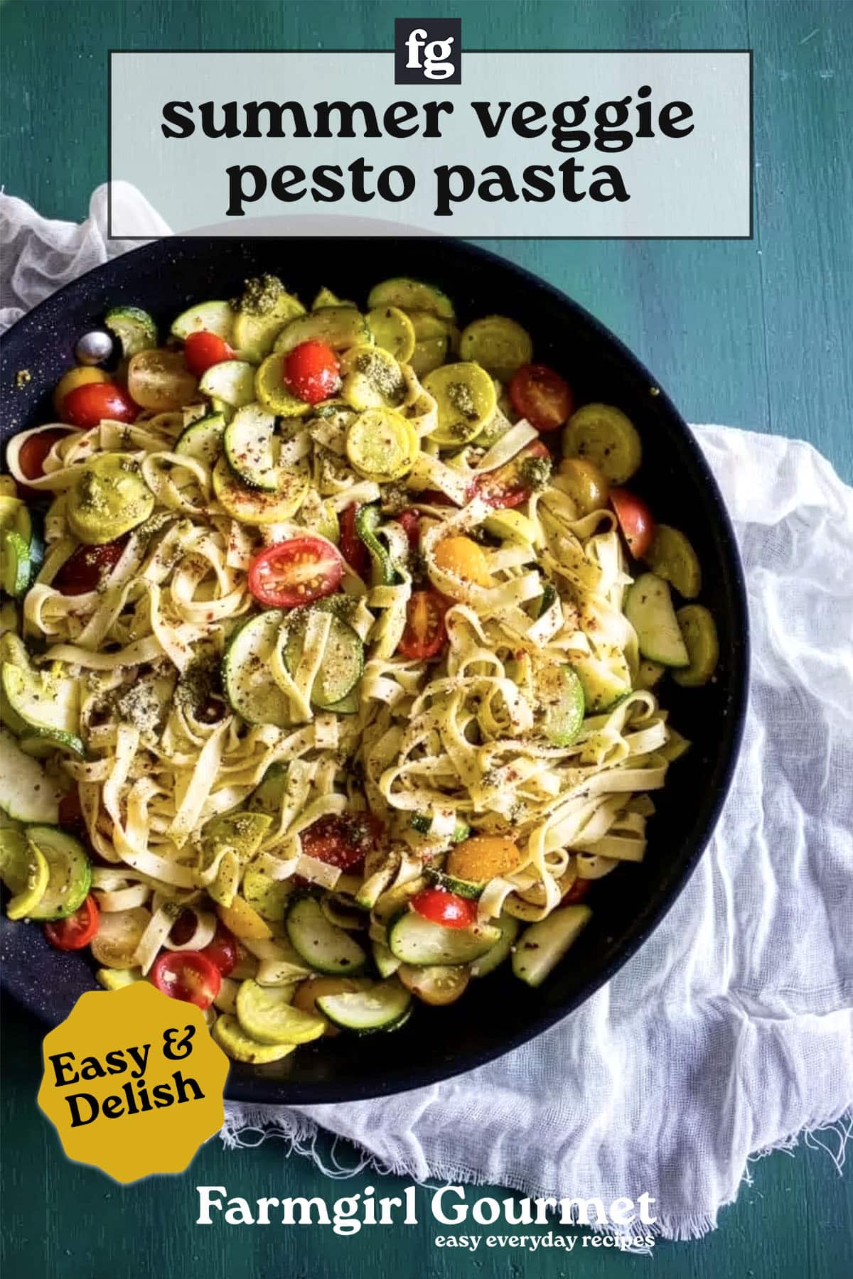 A skillet of summer vegetable pesto pasta with yellow squash, zucchini, cherry tomatoes, and herbs sits on a green table. Text reads “summer veggie pesto pasta”—a recipe from Farmgirl Gourmet.