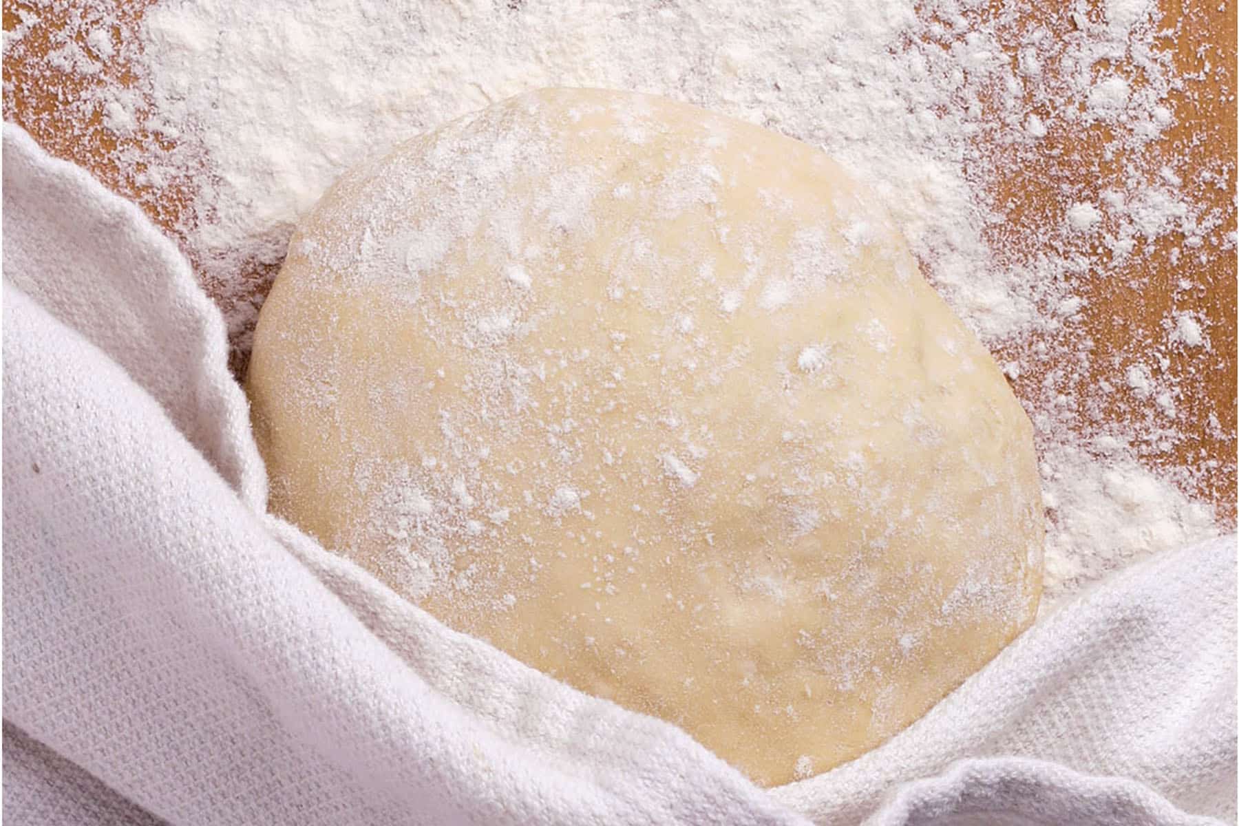 A ball of easy 20 minute pizza dough dusted with flour rests on a wooden surface, partially covered by a white kitchen towel, with flour scattered around.