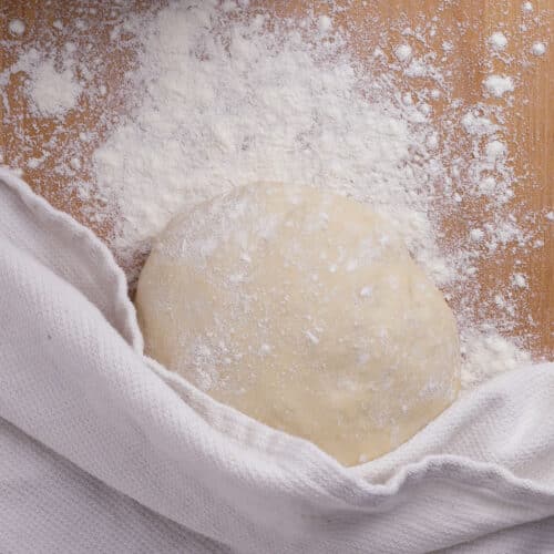 A ball of easy 20 minute pizza dough dusted with flour rests on a wooden surface, partially covered by a white cloth, with scattered flour and a glass bowl at the top of the image.