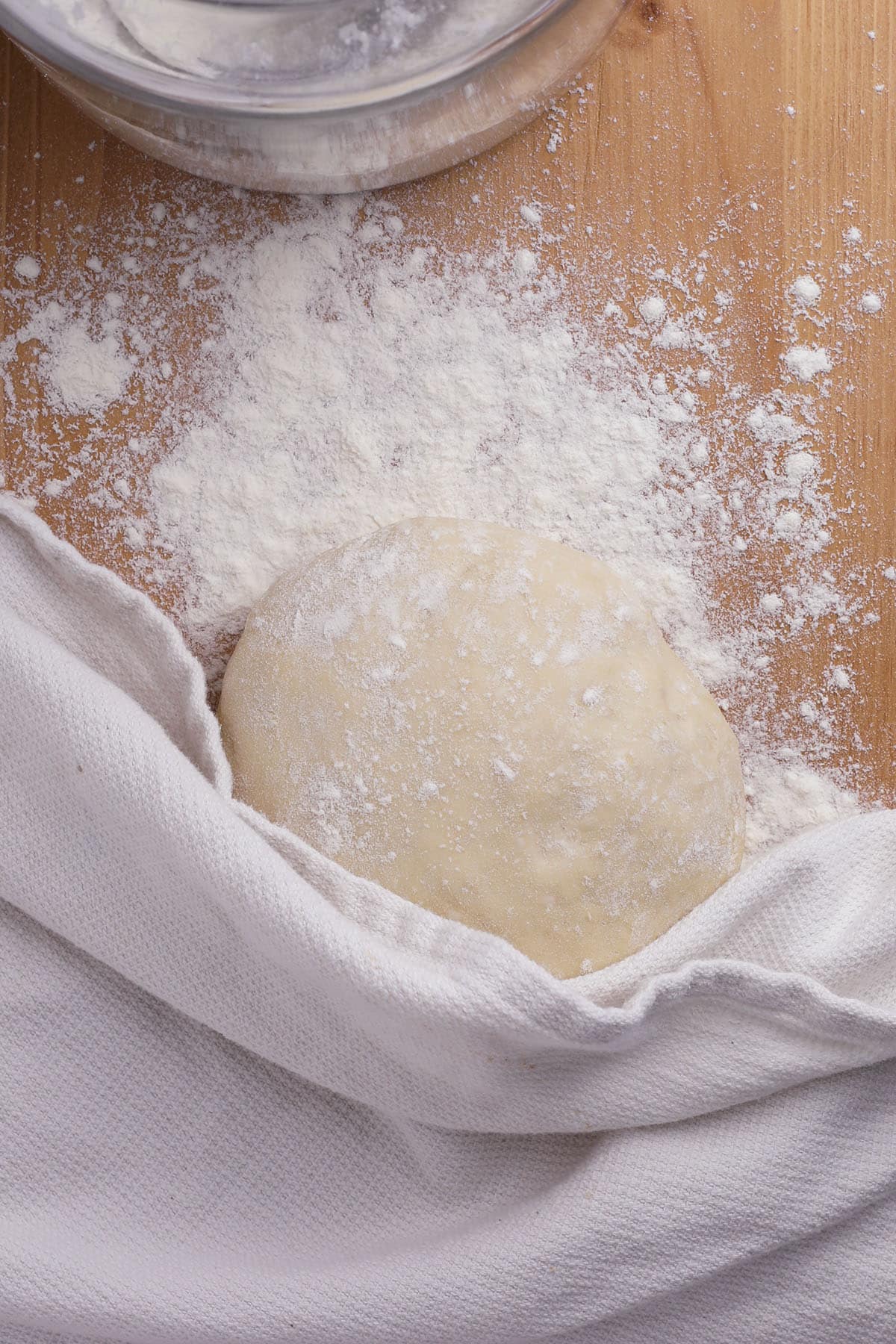 A ball of easy 20 minute pizza dough dusted with flour rests on a wooden surface, partially covered by a white cloth, with scattered flour and a glass bowl at the top of the image.