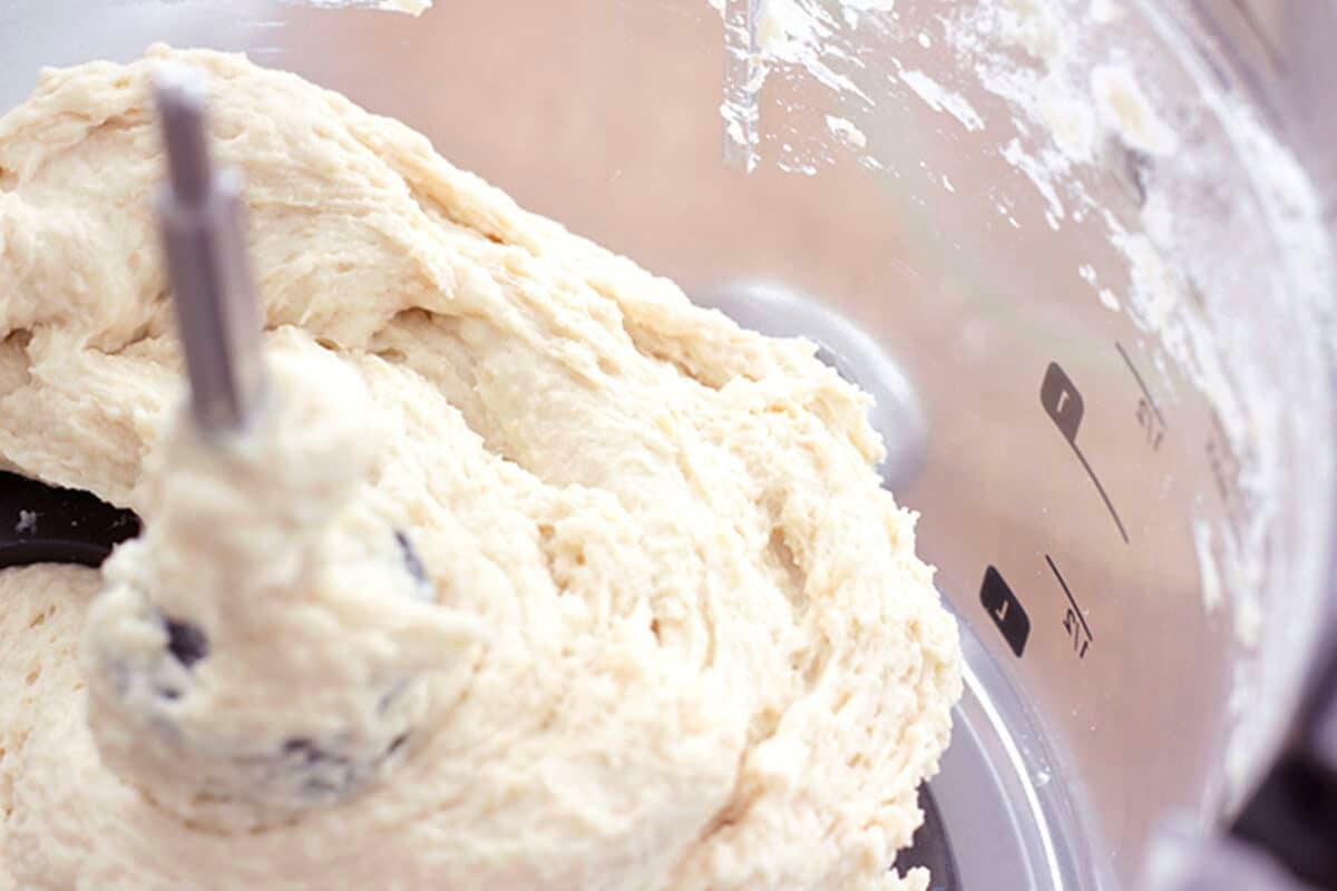 A close-up of thick, creamy easy 20 minute pizza dough being mixed in a clear food processor bowl, with dough clinging to the central blade and markings visible on the side of the bowl.