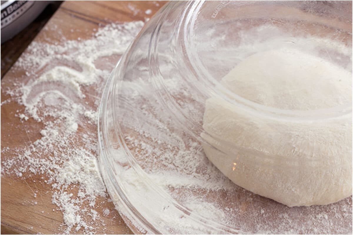 A ball of easy 20 minute pizza dough rests on a floured wooden surface, partially covered by an upside-down clear glass bowl. Flour is scattered around the dough.